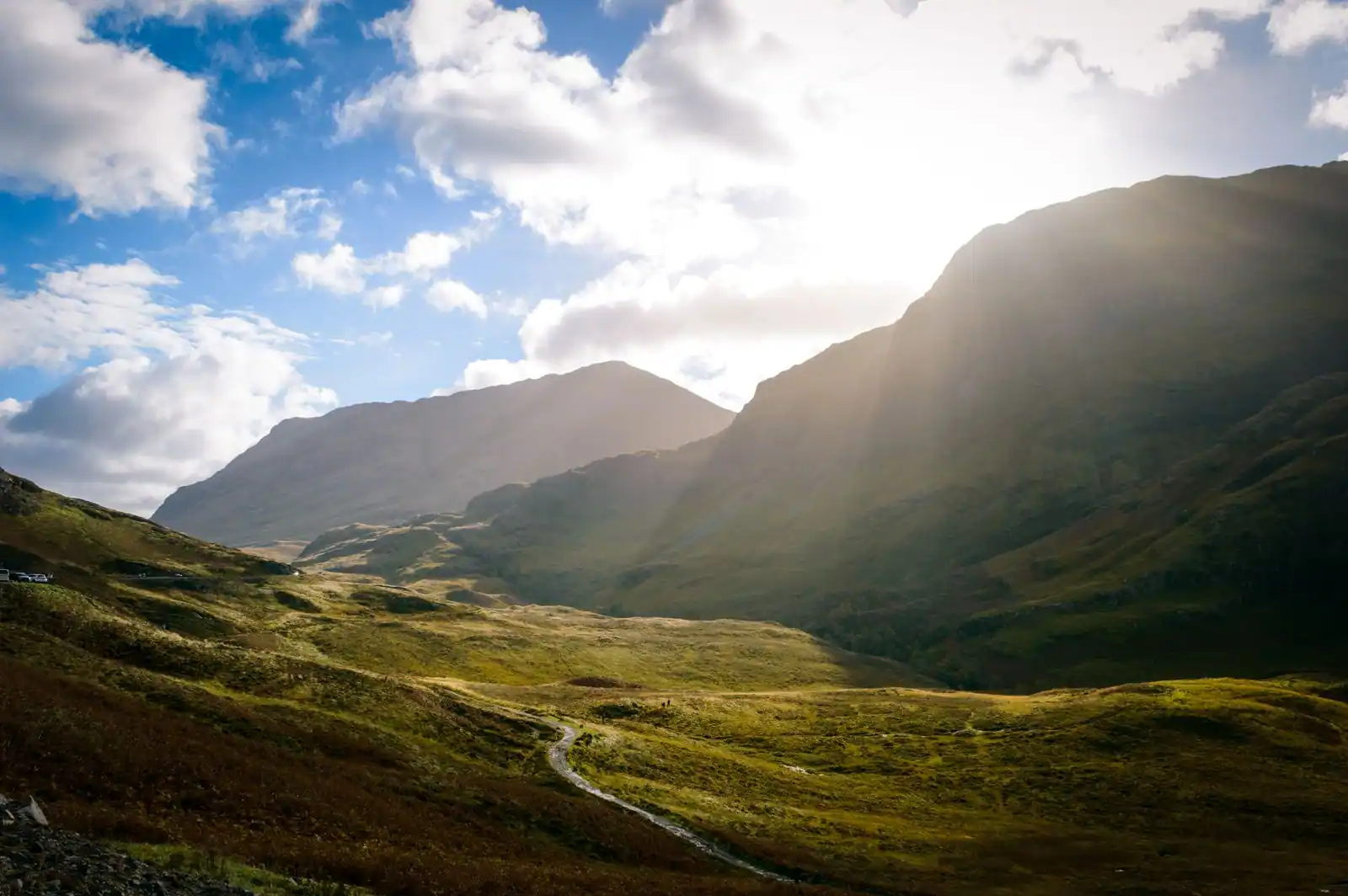 Vallée verdoyante des Highlands écossais entre des montagnes, traversée par un sentier sous un ciel ensoleillé