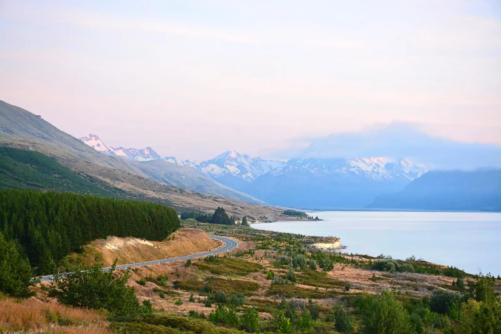 Lac et montagnes enneigées de Nouvelle-Zélande bordés d'une route sinueuse, sous un ciel pastel.