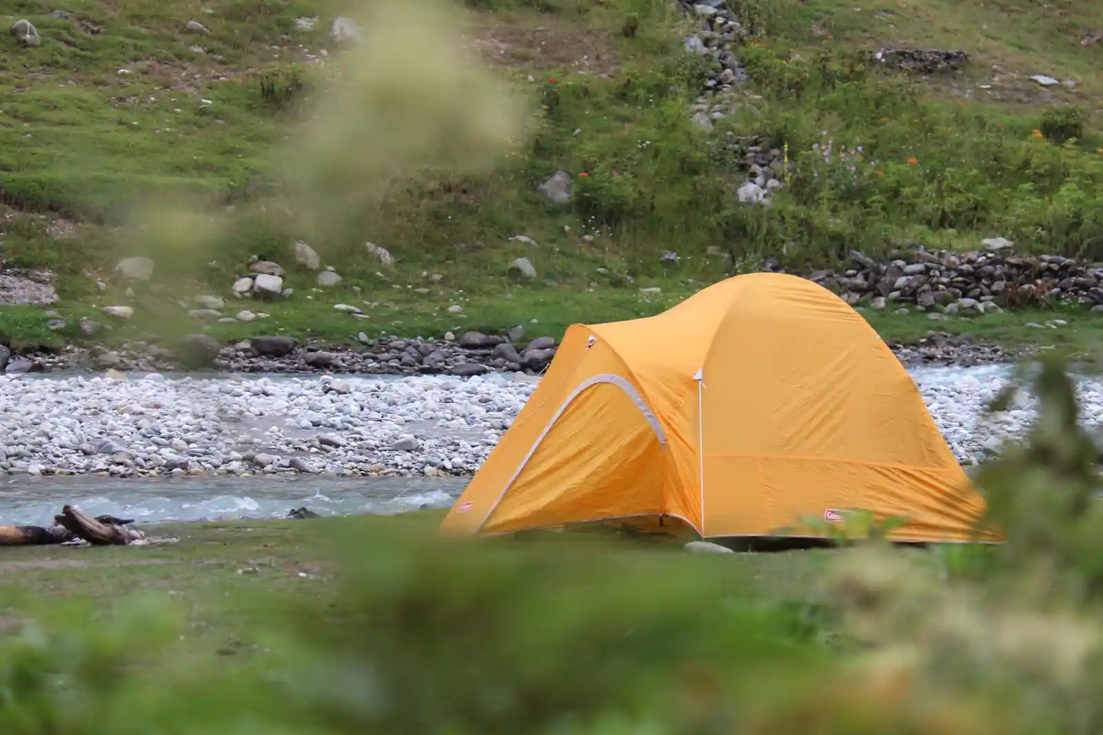 Tente orange plantée au bord d'une rivière de montagne, sur une berge herbeuse et un lit de galets.