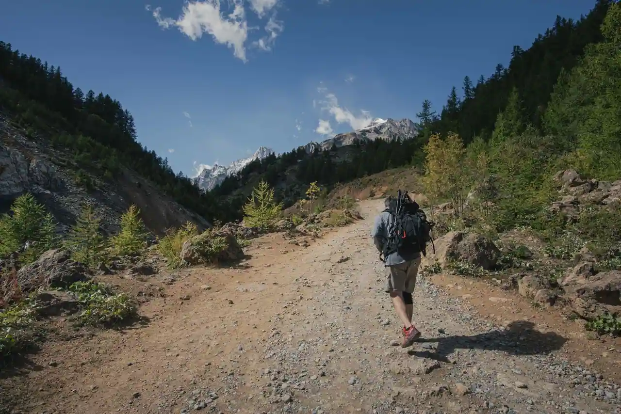 Randonneur au sac à dos de trek marchant sur un sentier de montagne, sommets enneigés au loin