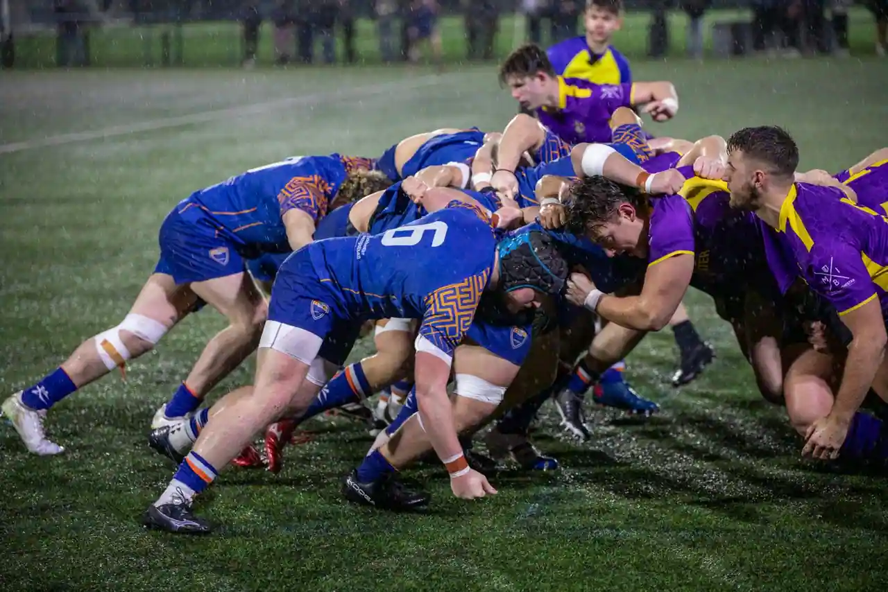 Mêlée de rugby sous la pluie lors d'un match nocturne, deux équipes à l'affrontement sur la pelouse.
