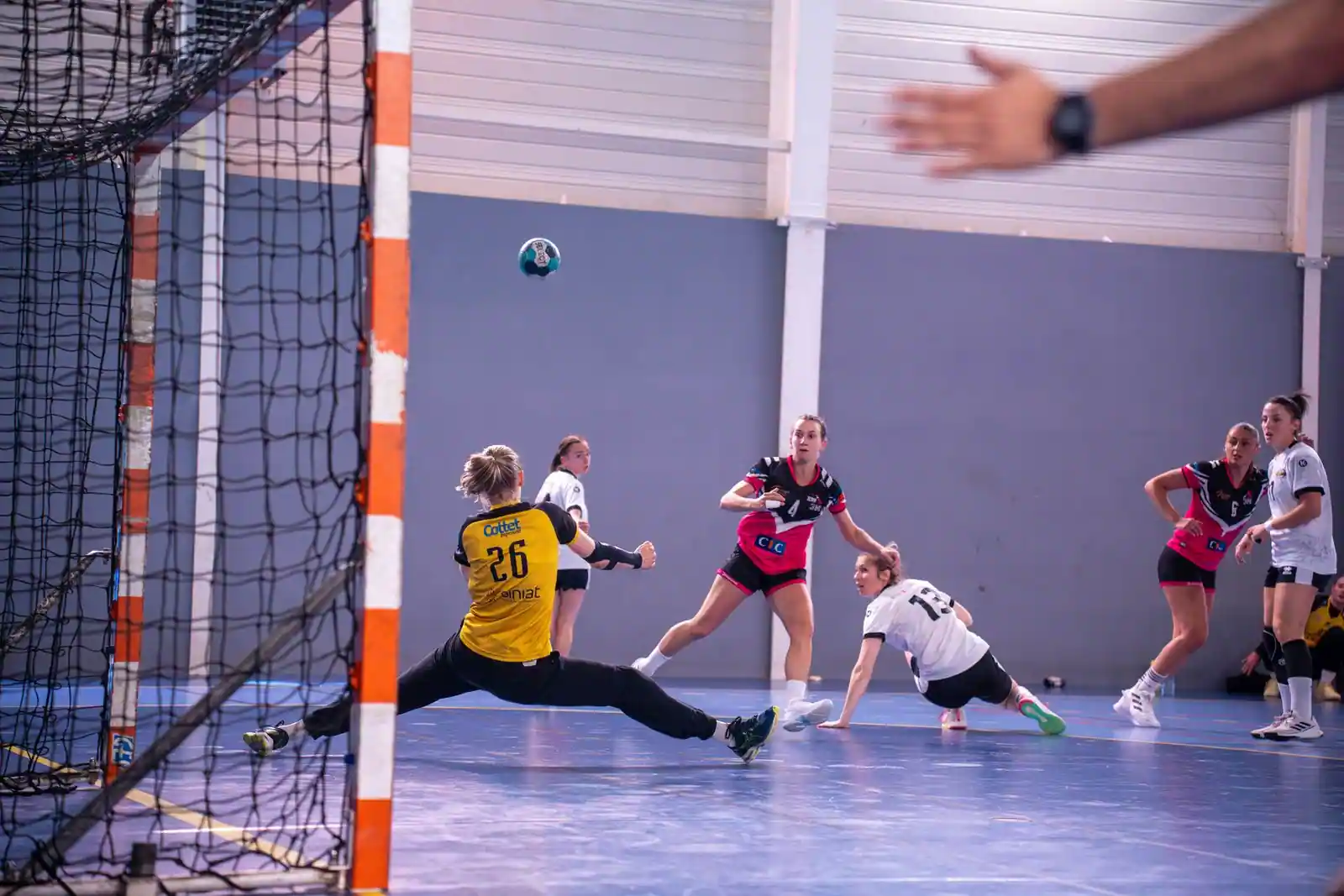 Match de handball féminin en salle : le ballon file vers la cage tandis que la gardienne plonge.