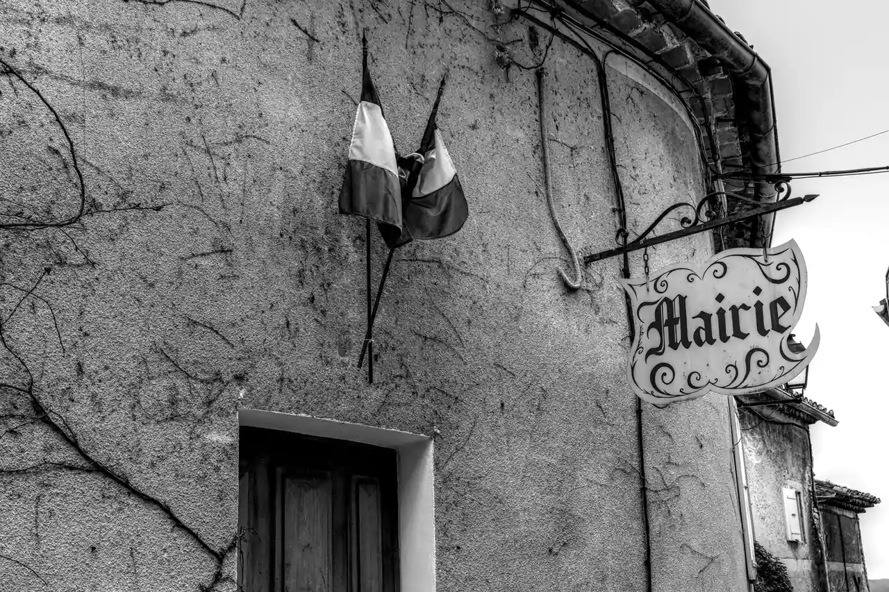 Enseigne « Mairie » et drapeaux français sur la façade d’une mairie de village, en noir et blanc.