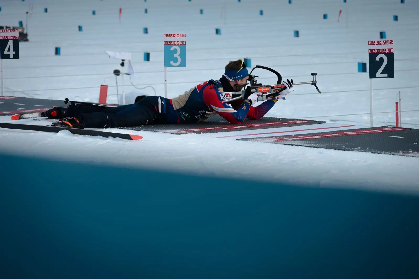 Biathlète en position de tir couché au stand, carabine épaulée face aux cibles, sur la neige.