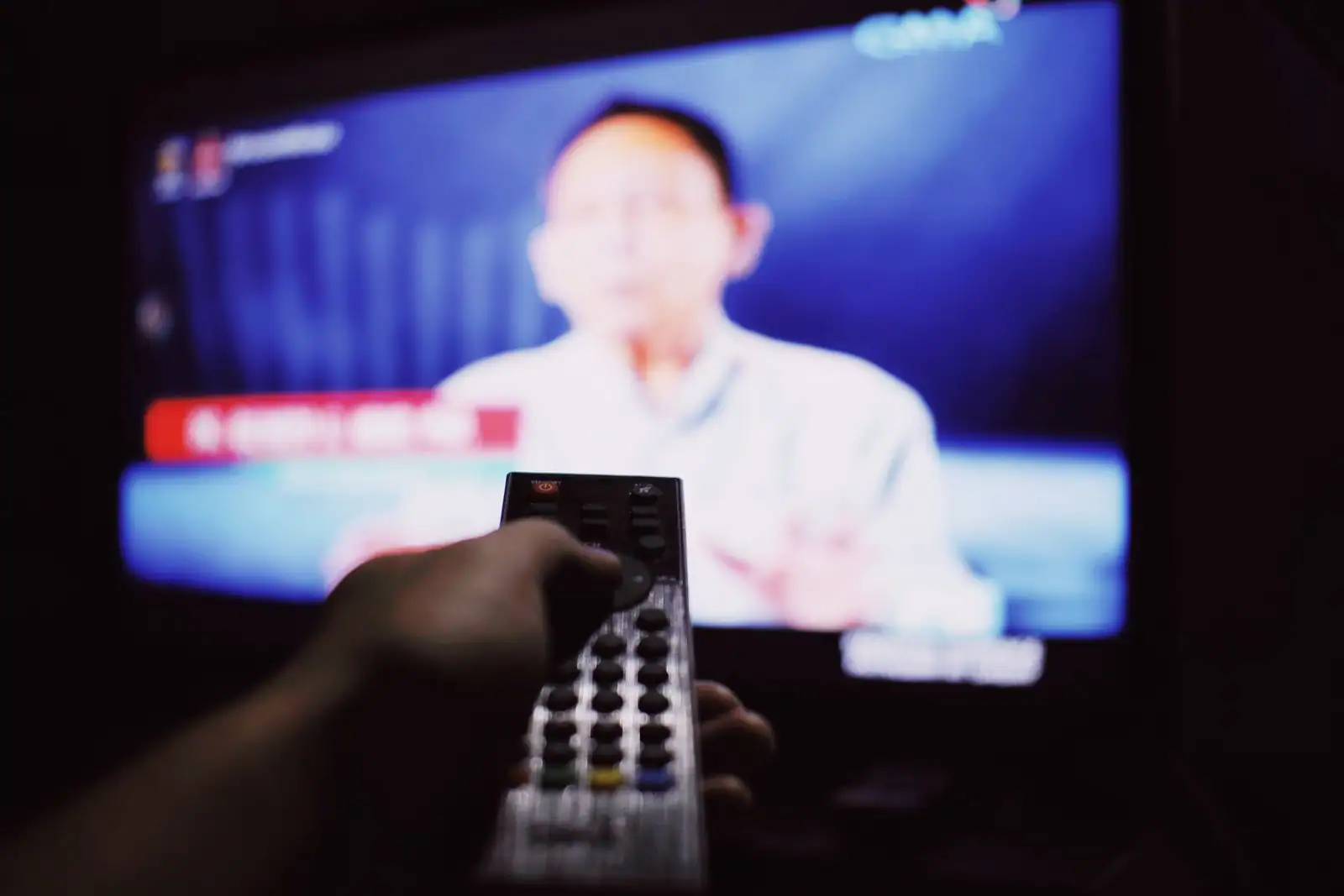 Main pointant une télécommande vers un téléviseur qui diffuse un programme, dans un salon