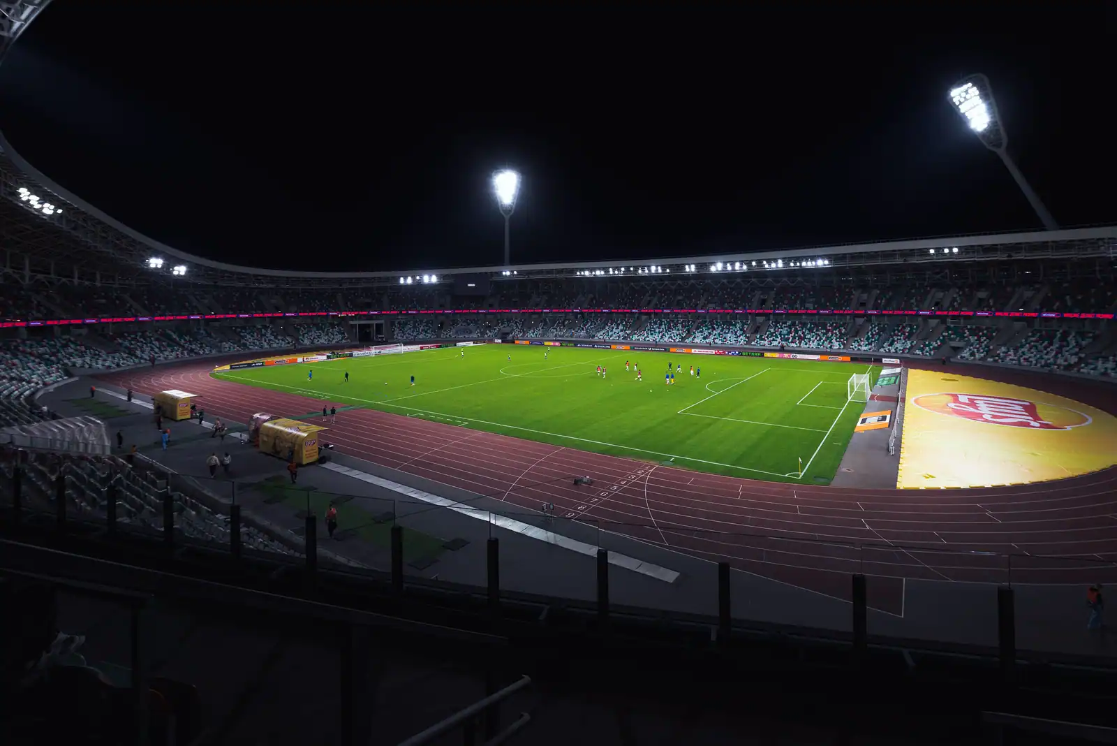 Stade de football illuminé la nuit, pelouse verte et gradins presque vides avant une rencontre.