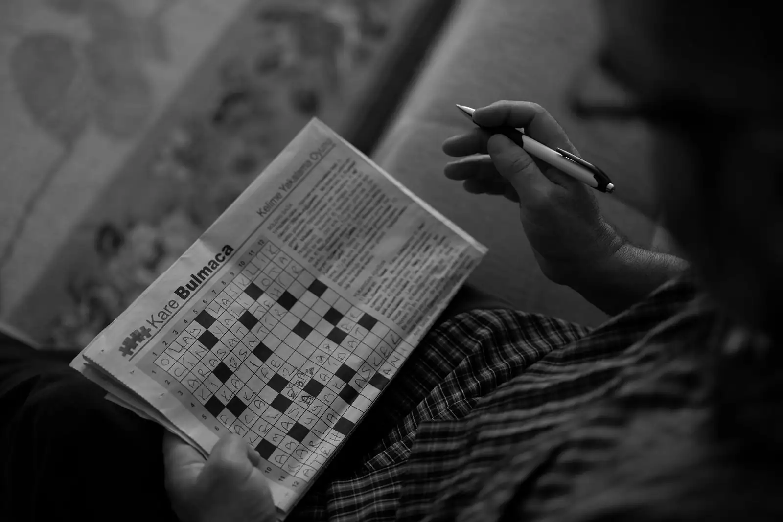 Personne remplissant au stylo une grille de mots croisés dans un journal, en noir et blanc
