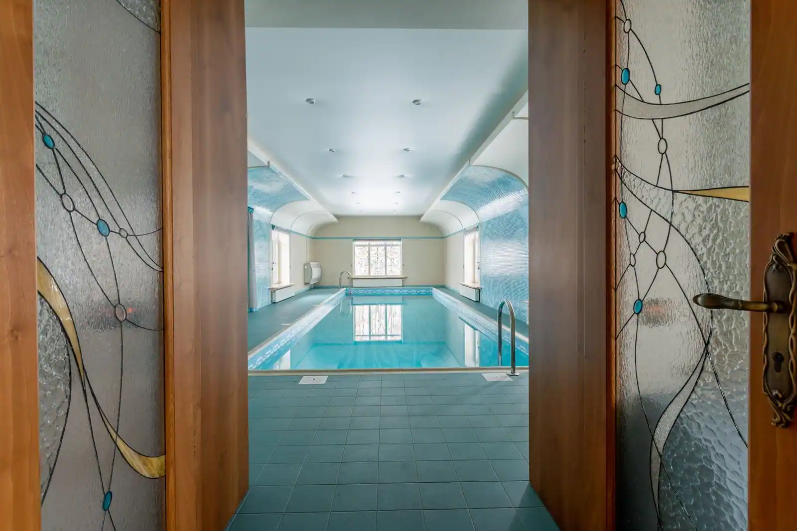 Piscine intérieure aux eaux turquoise, aperçue à travers une porte vitrée à motifs de vitrail
