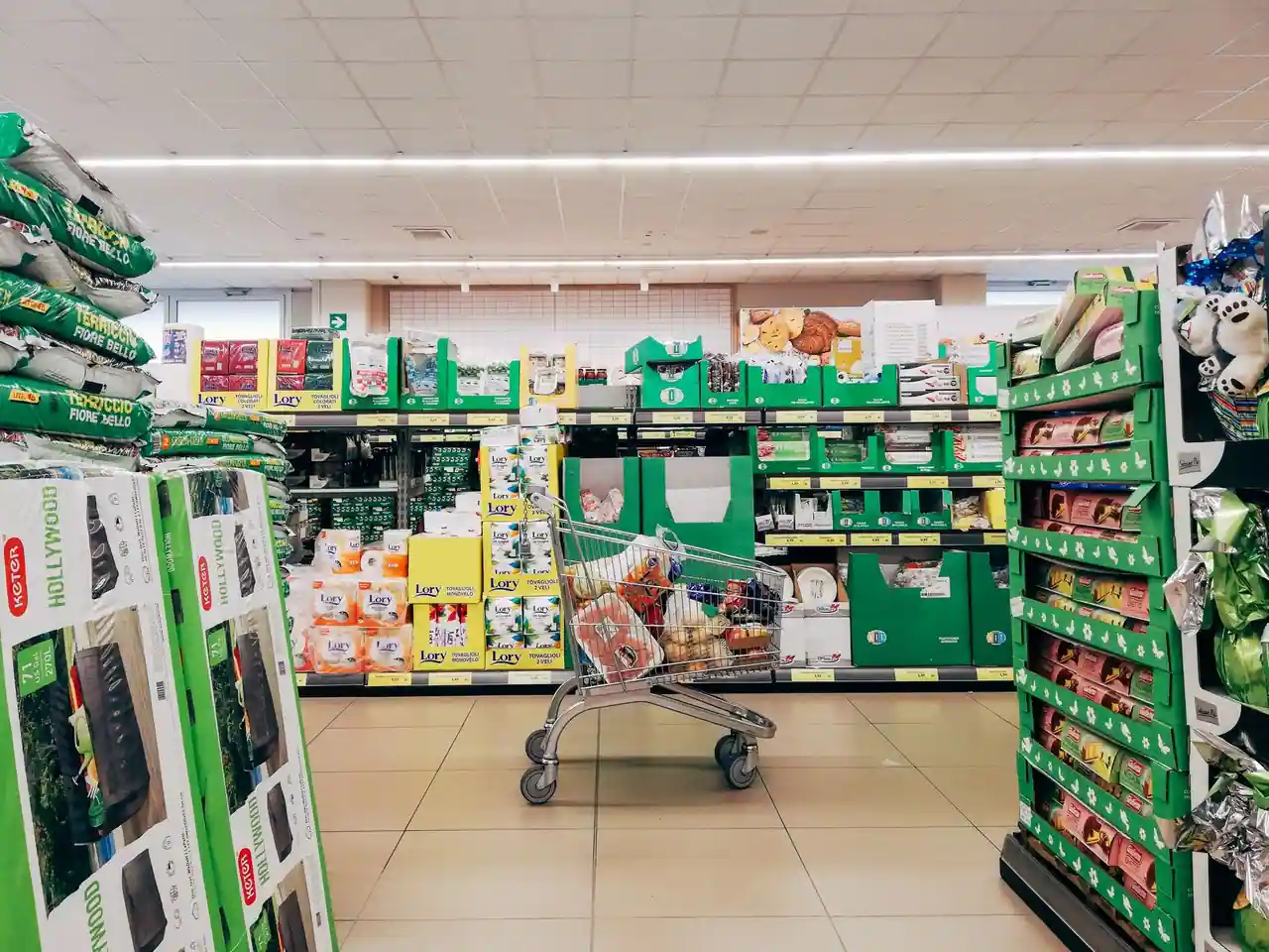 Chariot rempli de courses dans une allée de supermarché, avec étiquettes de prix jaunes en rayon.