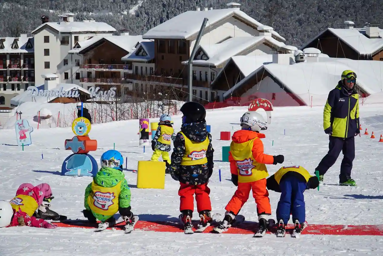 De jeunes enfants en cours de ski dans un jardin des neiges, encadrés par un moniteur, au pied des chalets enneigés.