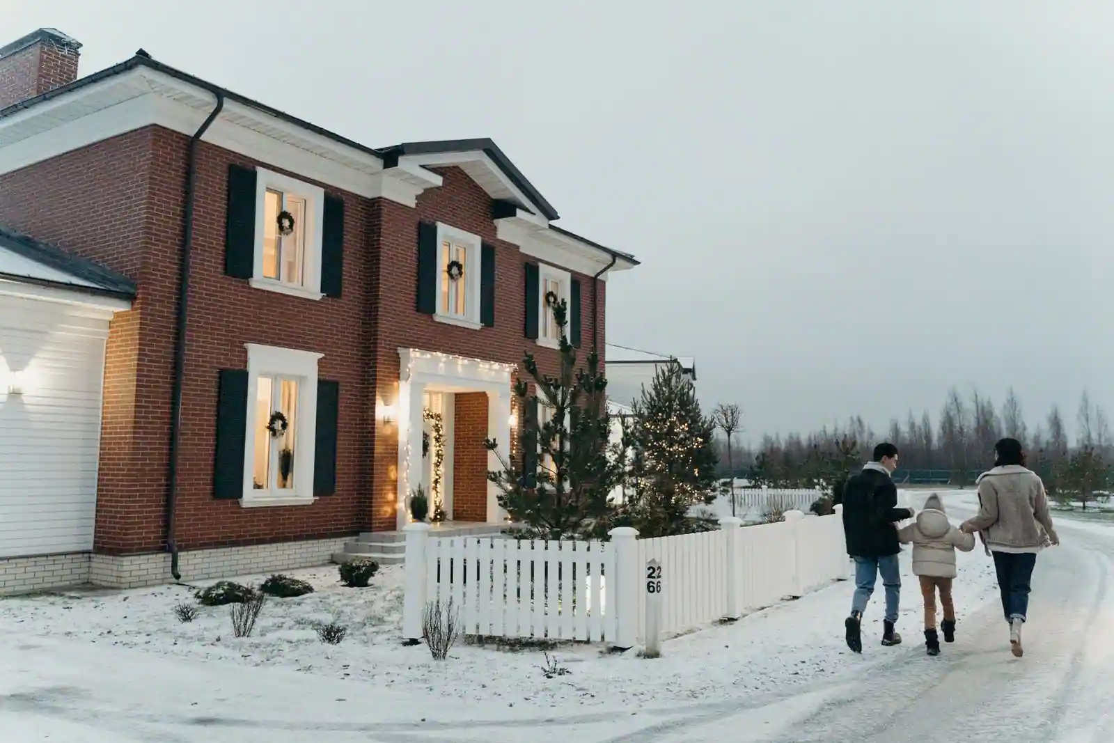 Famille marchant dans la neige devant une maison décorée pour Noël, guirlandes lumineuses et sapin.