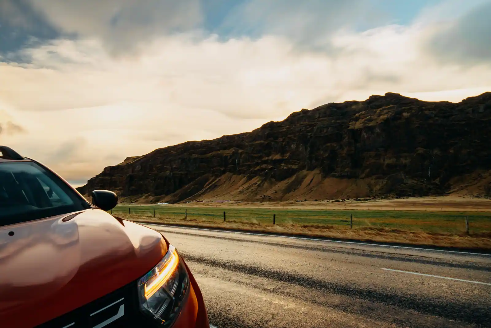 Voiture orange à l'arrêt au bord d'une route de campagne, devant un paysage de montagnes sous un ciel nuageux, ambiance de road trip.